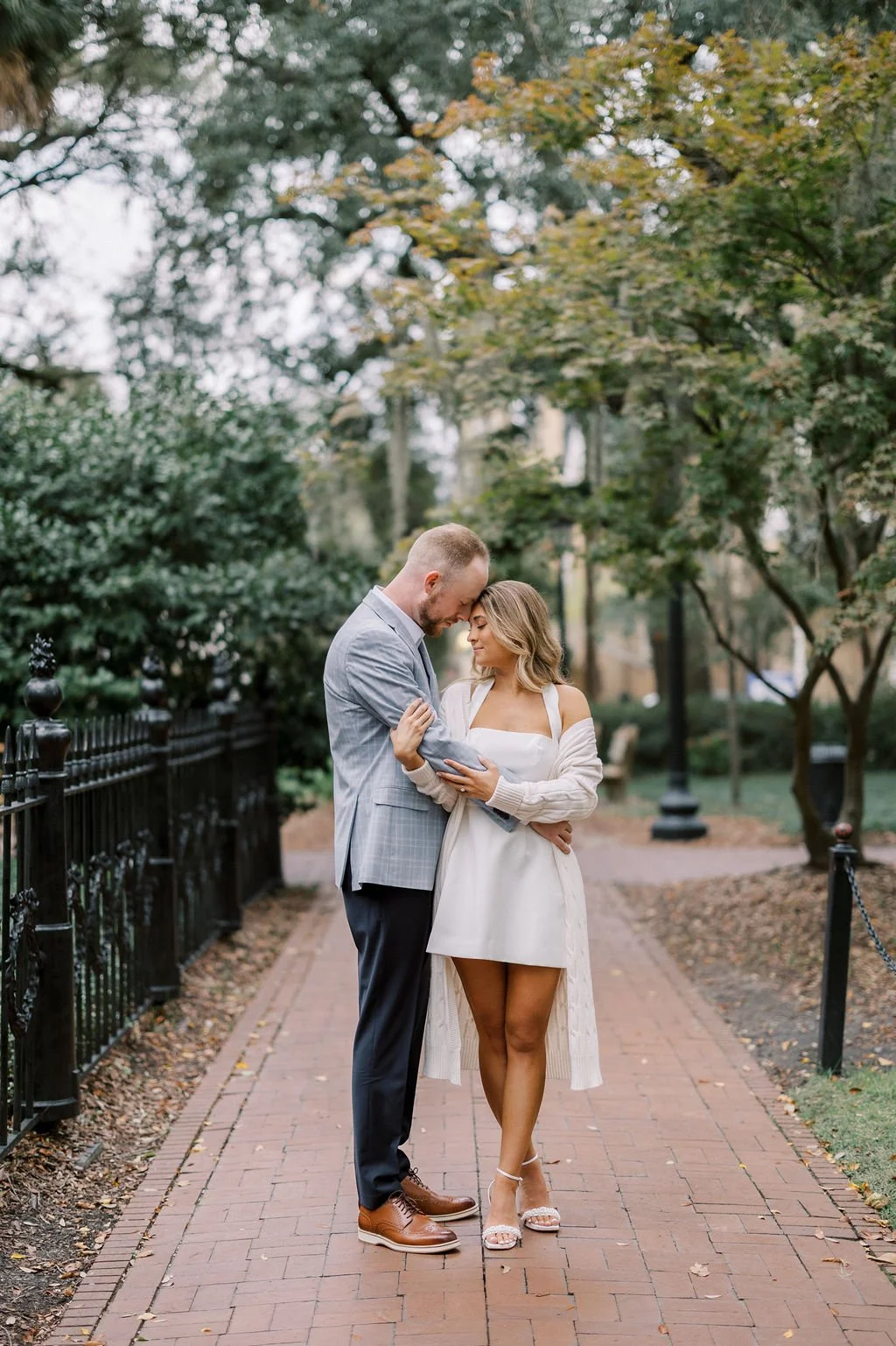 A couple sharing a tender moment on a brick path in a park with fall foliage. The man is wearing a gray blazer and dark trousers, and the woman is in a white dress with a long cardigan and high heels. They are touching foreheads and embracing.
