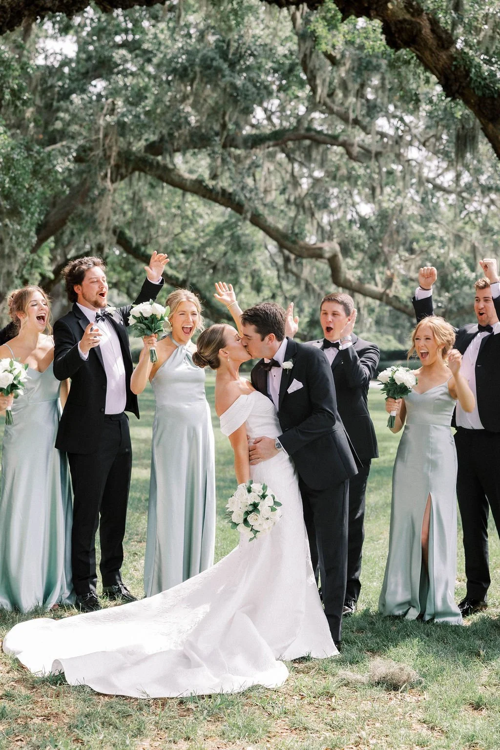 A wedding scene outdoors with a bride and groom kissing, surrounded by bridesmaids and groomsmen celebrating.