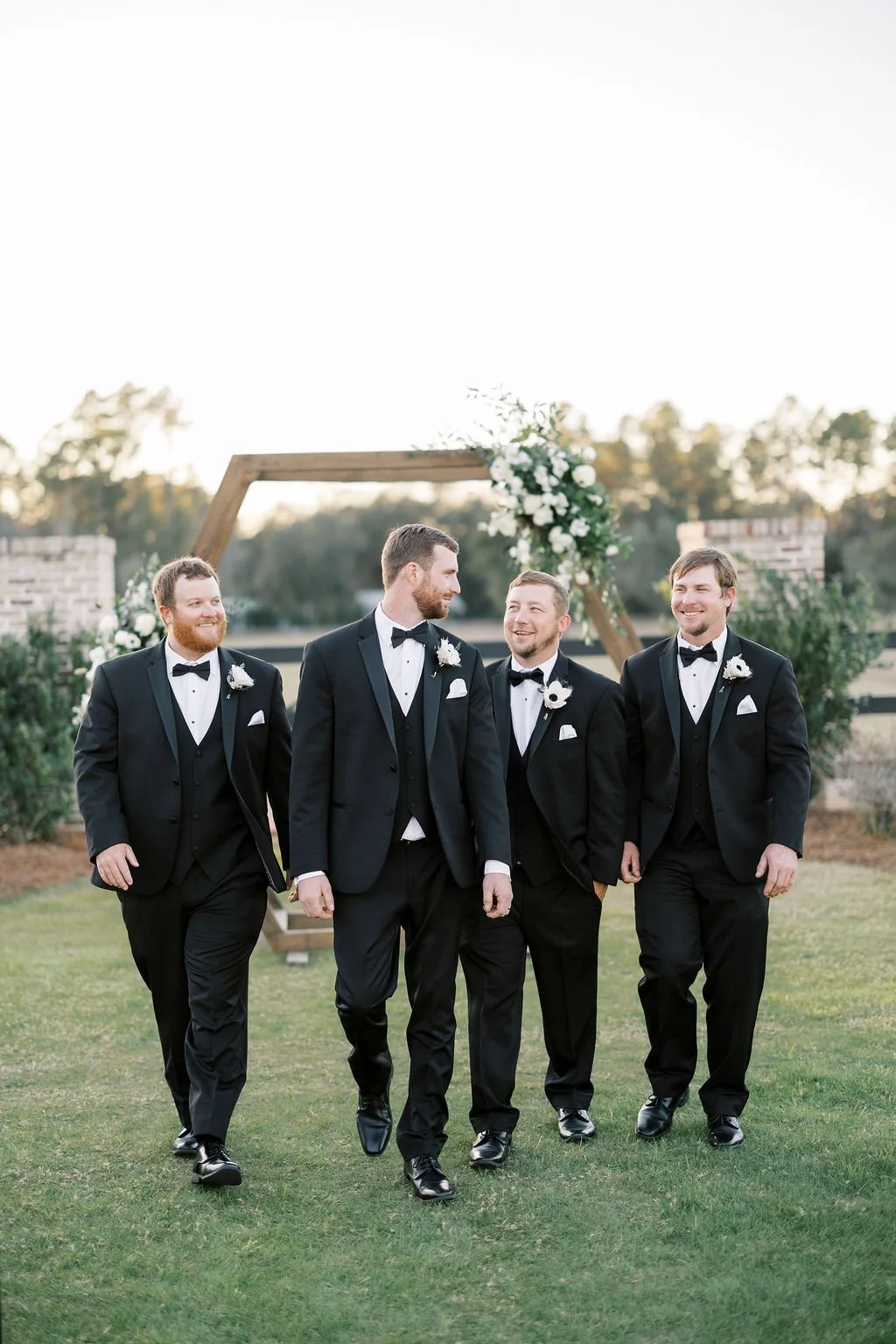 A group of five men dressed in black tuxedos with bow ties, walking outdoors on grass in front of a decorated wooden arch with white flowers, at a wedding or formal event during daytime.