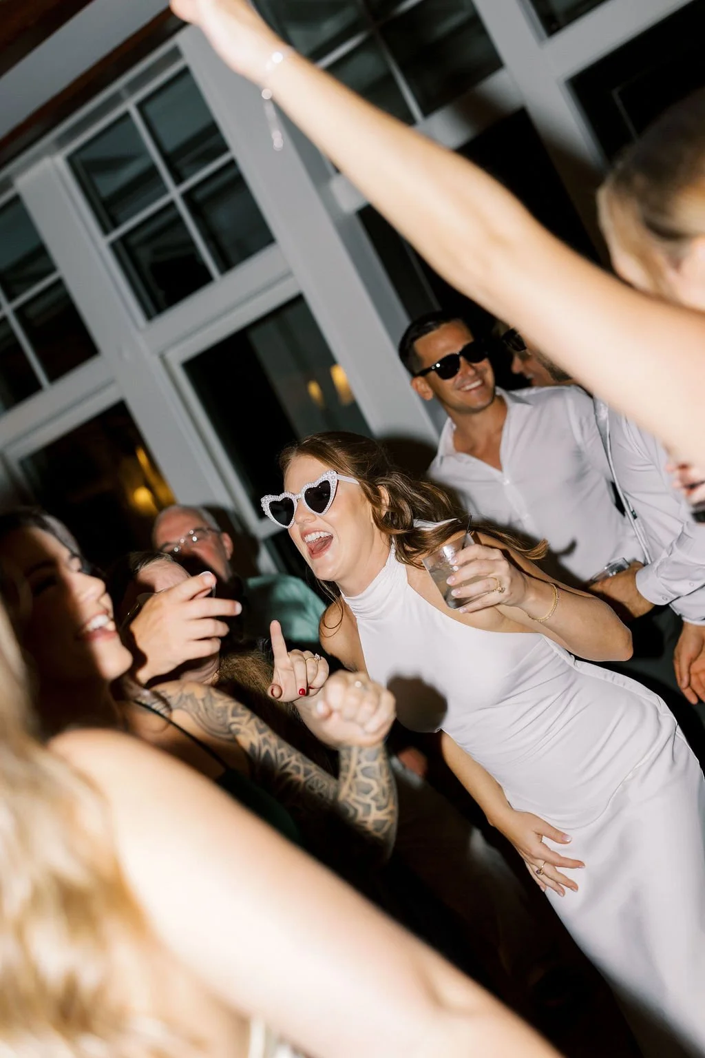 Group of people dancing and having fun at party, woman with heart-shaped sunglasses, others with sunglasses, smiling, holding drinks, lively atmosphere