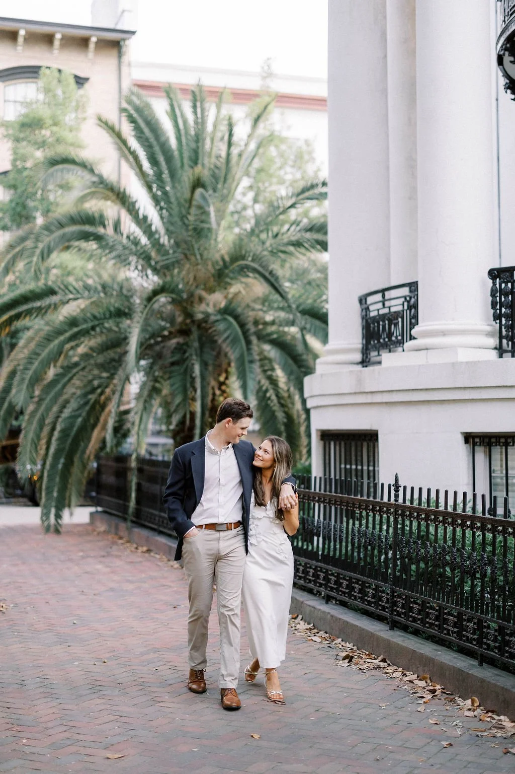 A young couple walking arm in arm on a brick sidewalk, smiling and looking at each other, with a large palm tree and a historic white building with black wrought iron fences in the background.