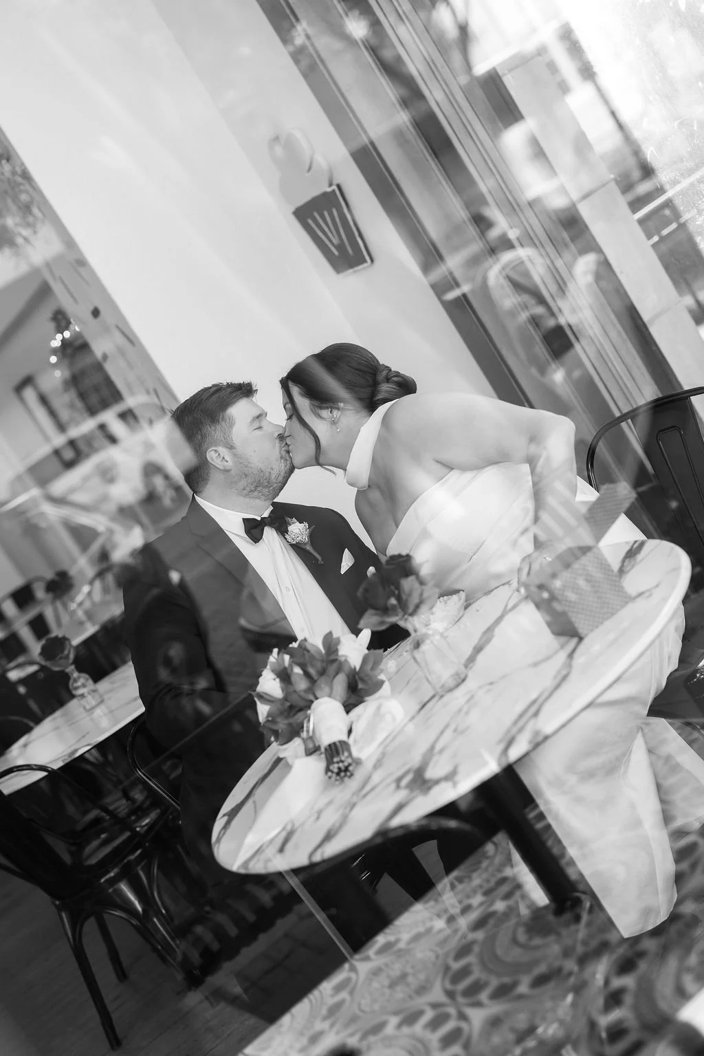 A black and white photo of a newlywed couple sharing a kiss inside a restaurant or cafe with large windows, reflected trees, and tables with floral arrangements.