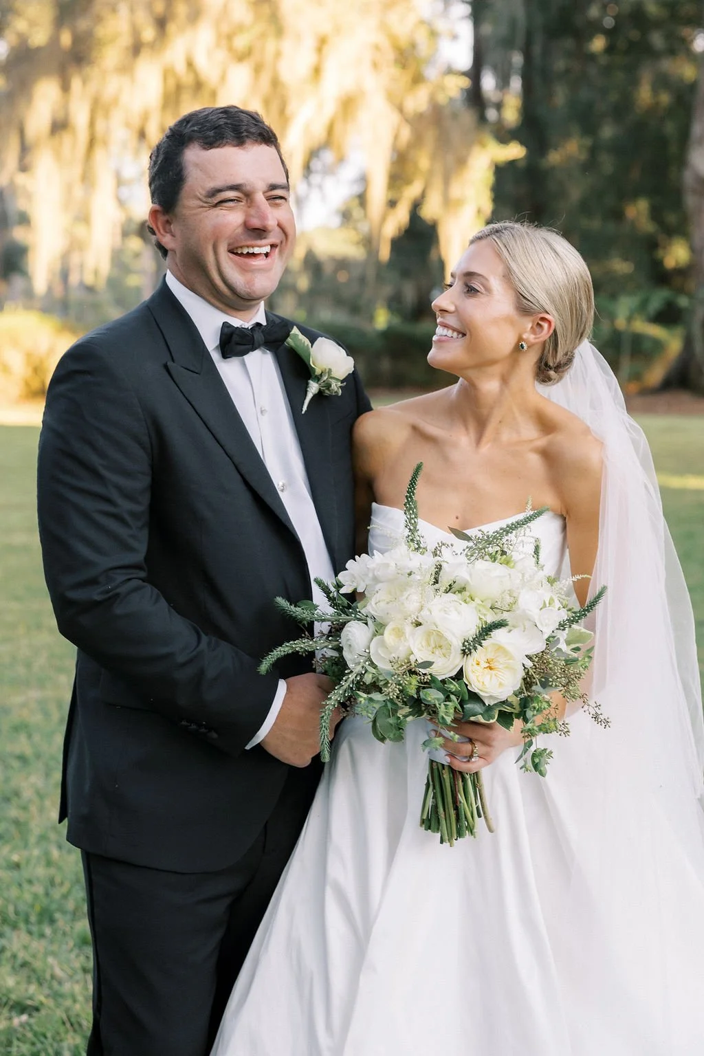 A groom in a black tuxedo and a bride in a white wedding dress standing outdoors, smiling at each other. The bride holds a large bouquet of white flowers and greenery, and they are surrounded by trees and sunlight.