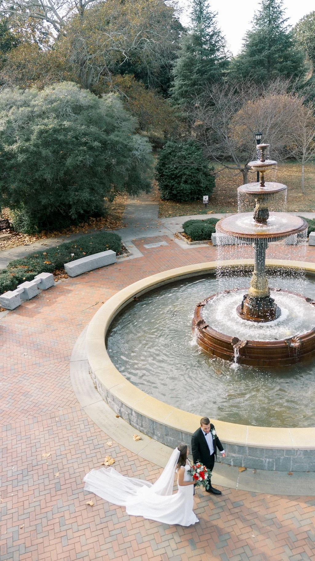 A bride and groom walking near a fountain outdoors on a brick plaza, with the bride holding a bouquet of red and white flowers and wearing a long white gown while the groom is in a black tuxedo.
