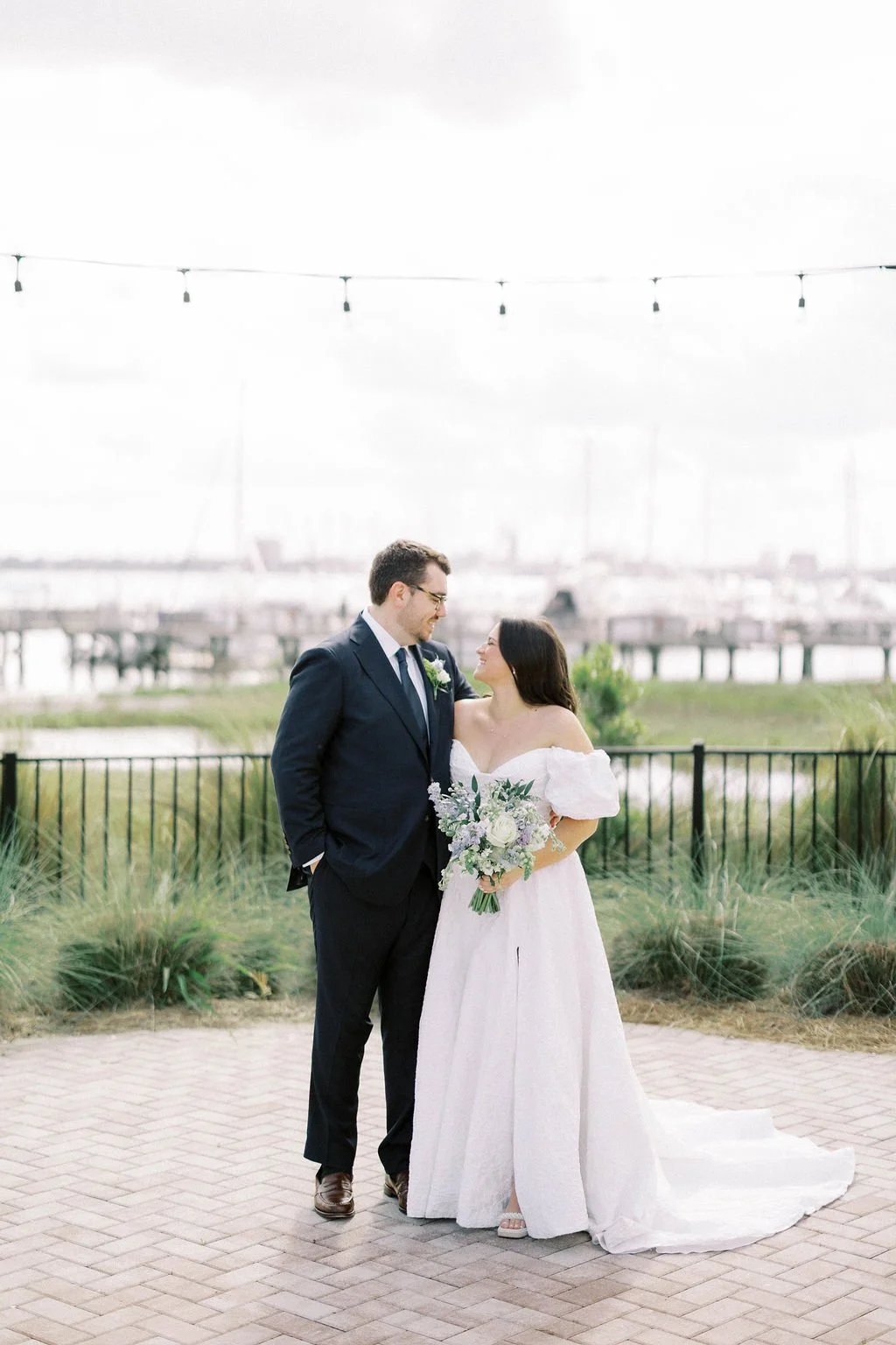 A bride and groom standing outdoors on a paved area, with water and a marina in the background. The bride is holding a bouquet and wearing a white off-shoulder wedding dress, while the groom is dressed in a dark suit and tie. They are smiling and loo