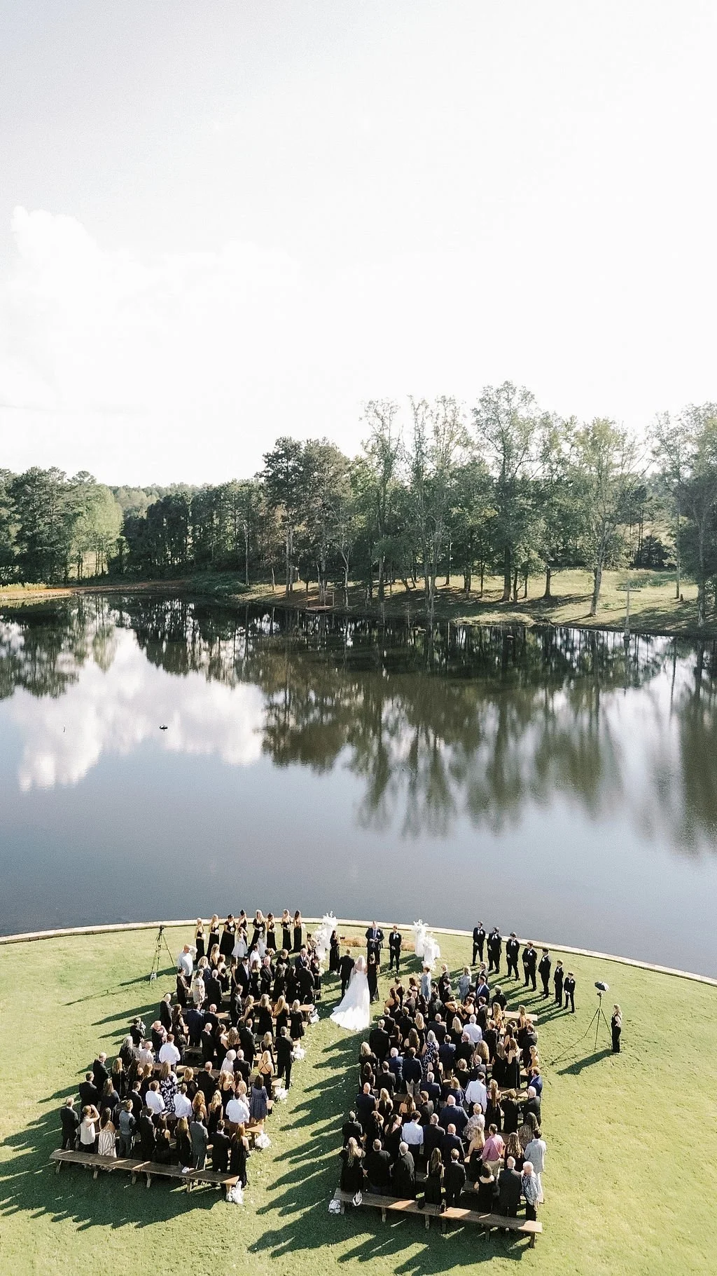 An outdoor wedding ceremony taking place on a green lawn near a lake, with guests seated and standing, a bride and groom at the altar, and trees reflected in the water.