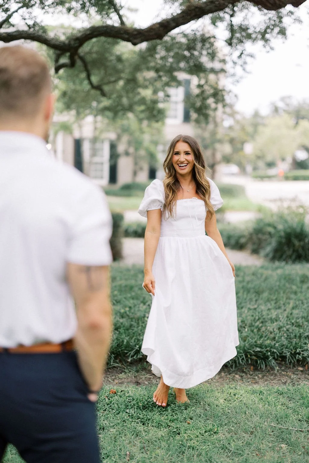 A woman in a white dress smiling and looking at a man, who is partially visible in the foreground, outdoors in a park-like setting.