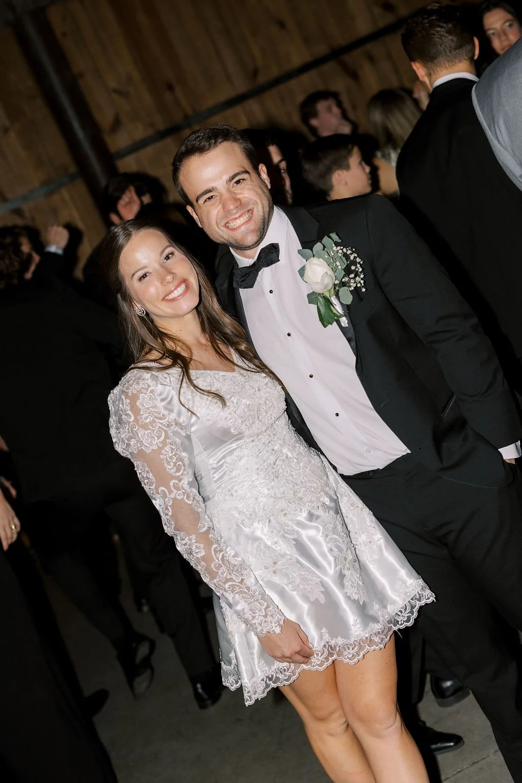 A smiling bride in a white lace dress and a smiling groom in a tuxedo with a white boutonniere, at a wedding reception with guests in the background.