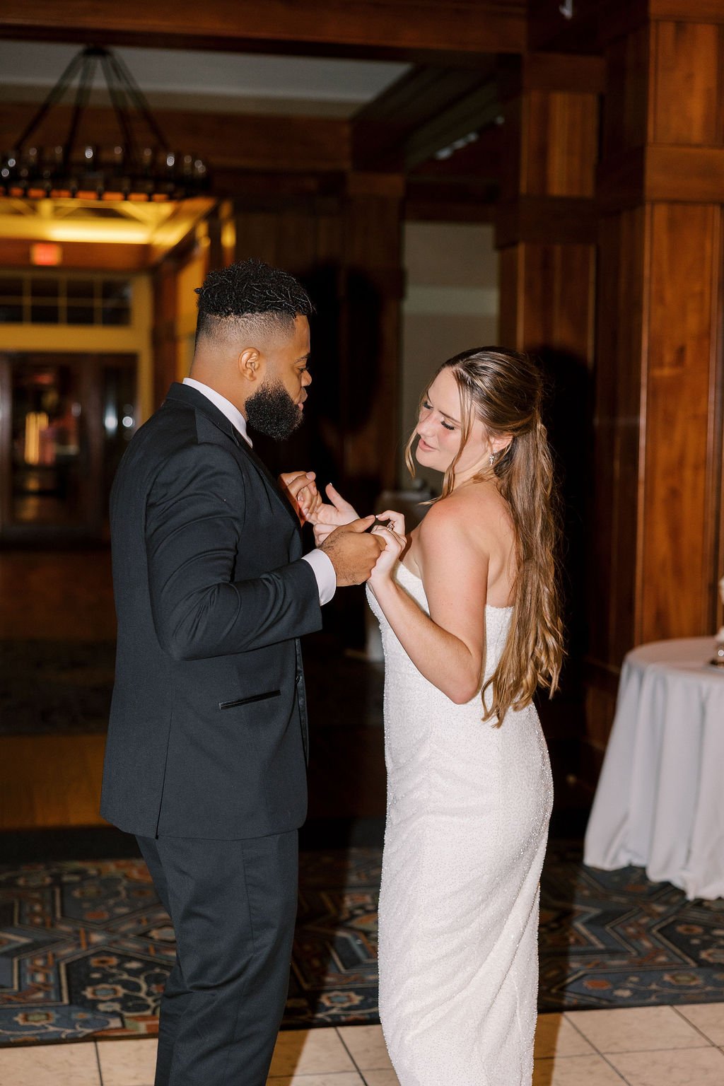 A bride and groom dancing during their wedding reception in a warmly lit wooden venue, with the groom in a black suit and the bride in a strapless white wedding dress.