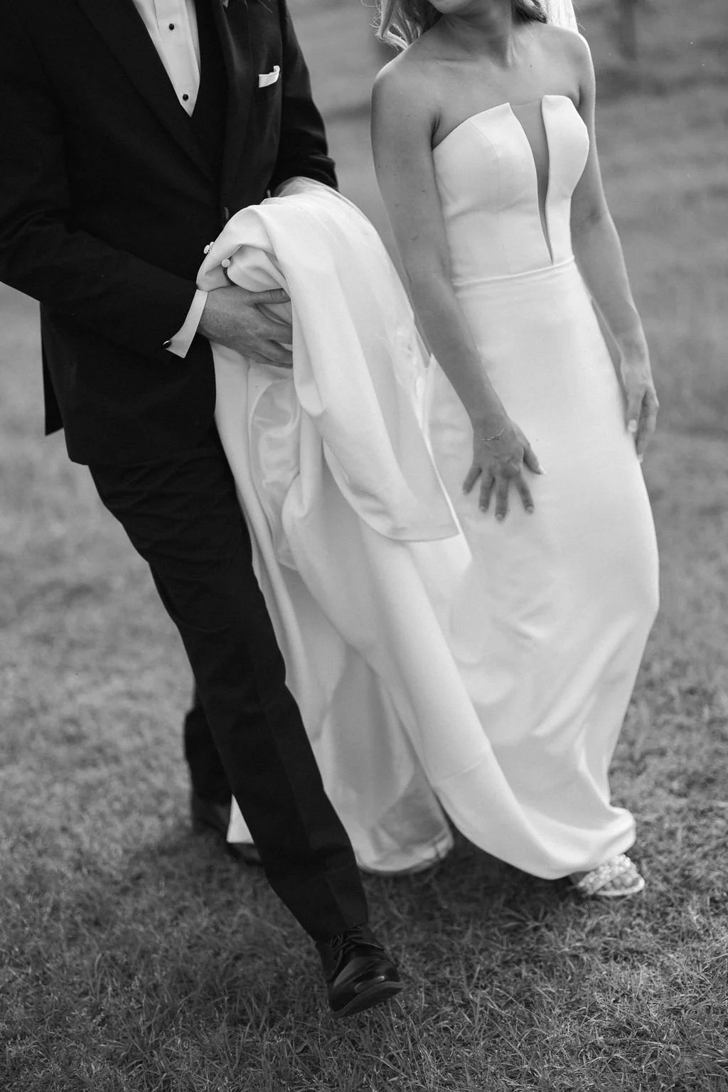Bride and groom walking outdoors, groom holding the bride's dress, black and white photo.