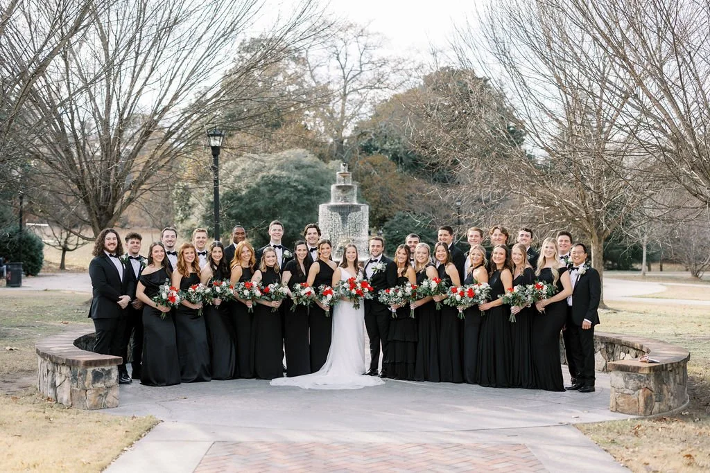 A large wedding party dressed in formal black attire, posing outdoors in front of a fountain, with a bride in a white gown and a groom in a black tuxedo at the center, holding bouquets of red, white, and green flowers.