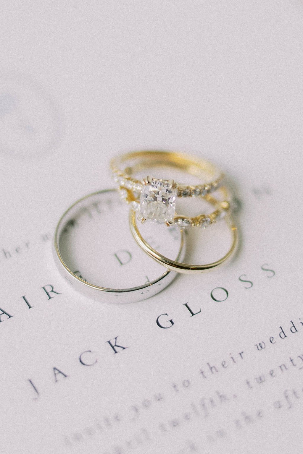 Close-up of three gold rings, one featuring a large diamond, resting on a wedding invitation with partially visible text.
