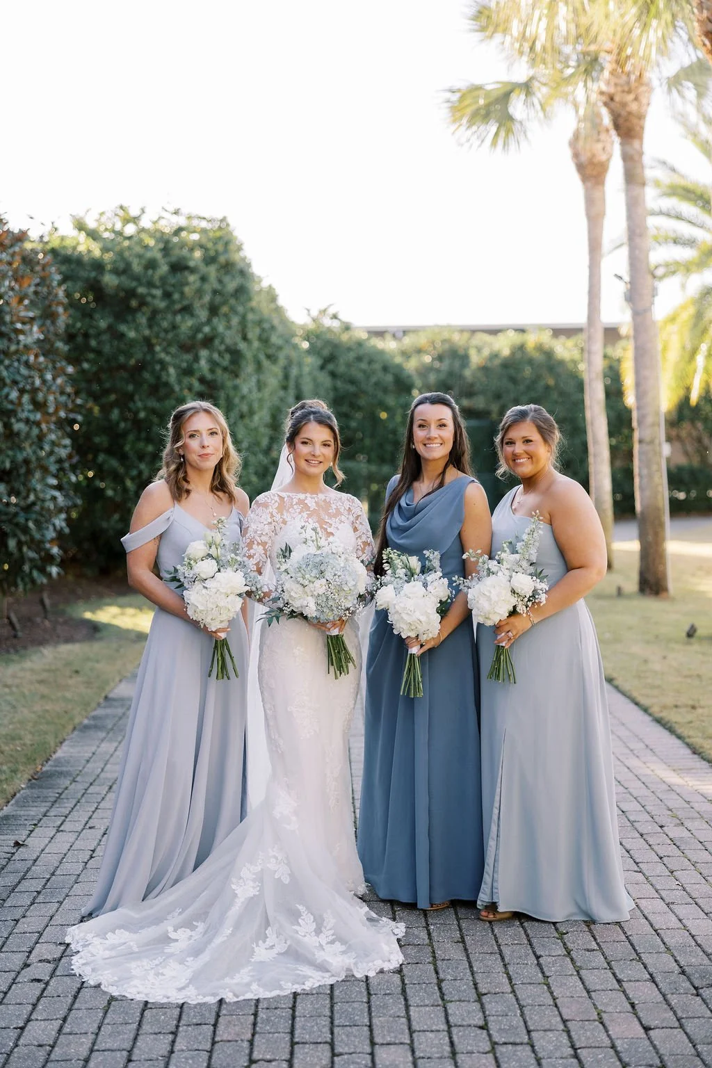 Bridal party standing on a stone pathway outdoors, holding bouquets of white flowers, with greenery and palm trees in the background, during daytime