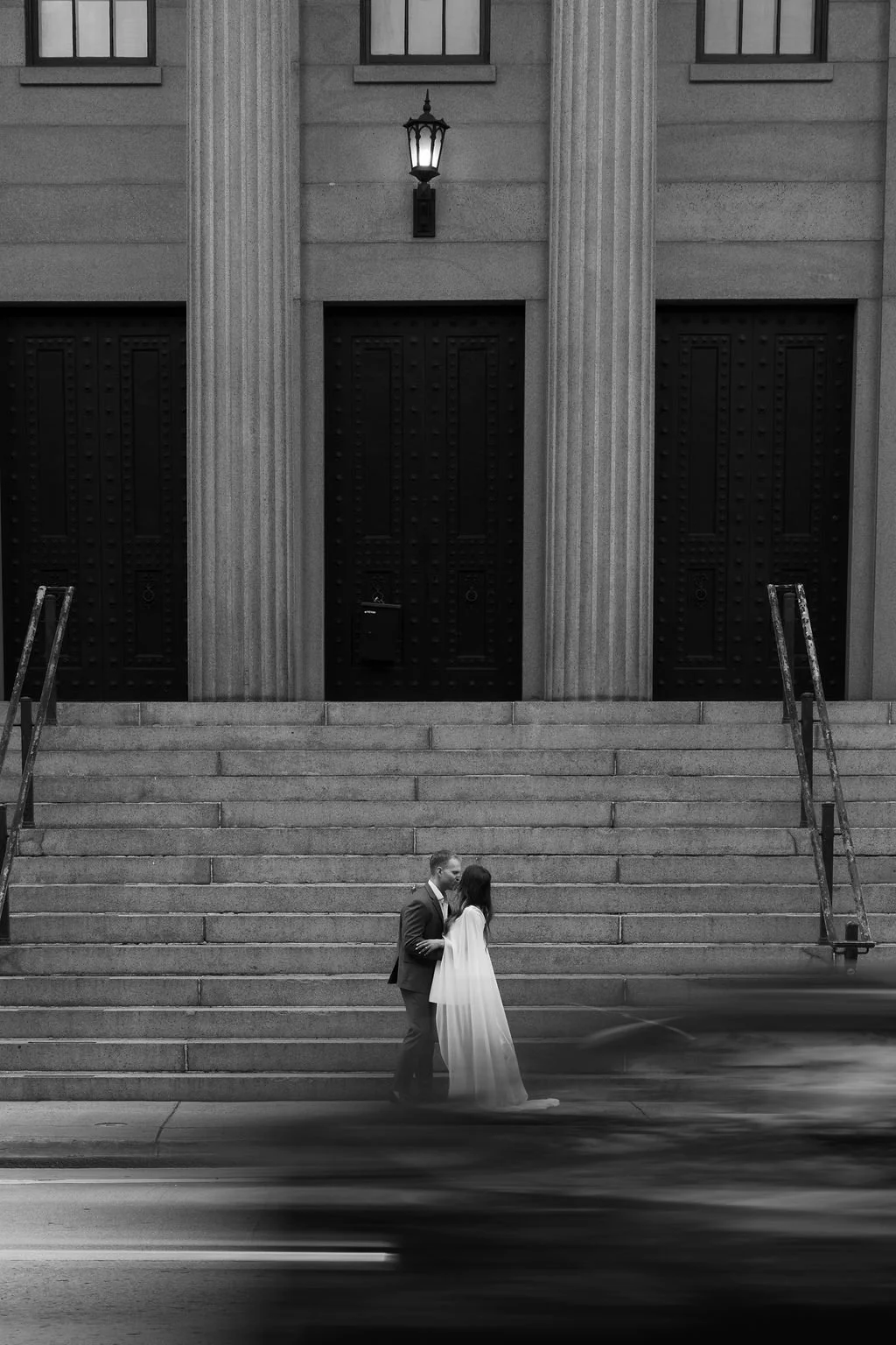 A bride and groom share a kiss on the steps of a large, classical building with tall columns and black doors, a street scene with a blurred car passing by in motion.