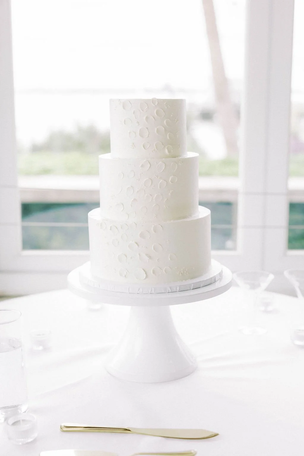 White three-tiered wedding cake with subtle circular pattern, placed on a white cake stand near large windows with sheer curtains, with utensils and glasses on the table.