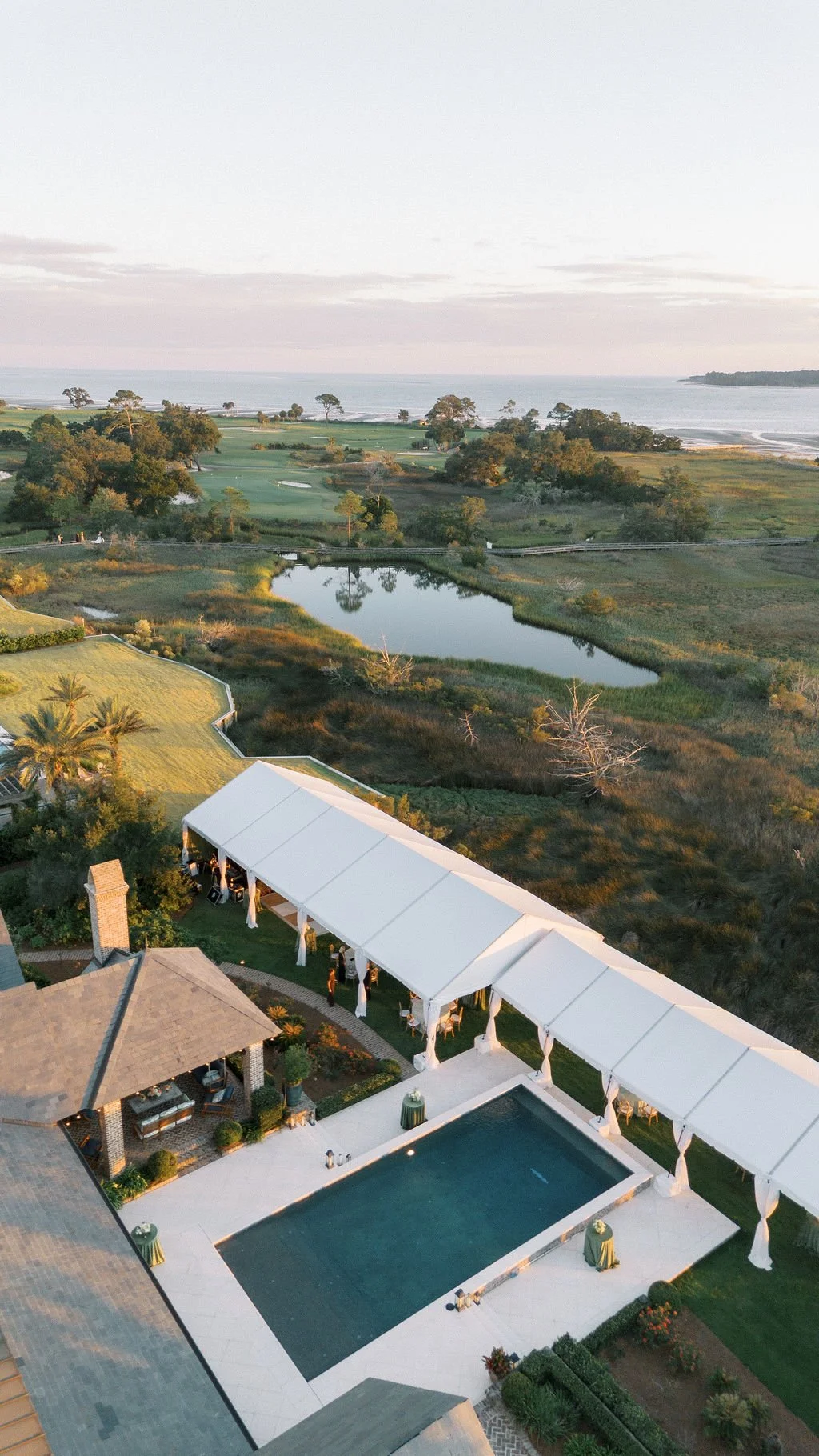 An outdoor event setup with a pool, covered seating area, and scenic view of lush greenery, a lake, and the ocean in the background during sunset.
