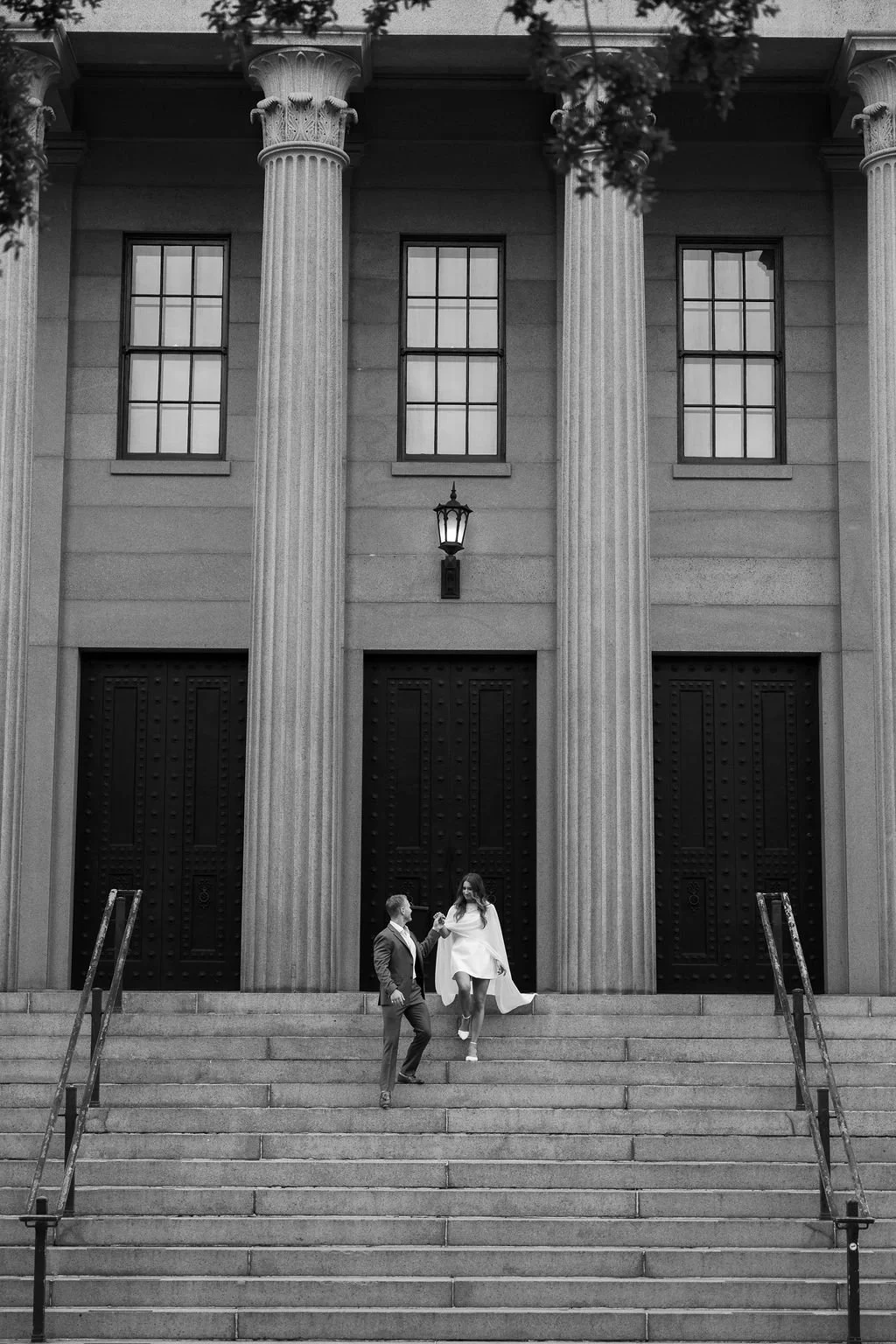 A man and woman on staircase in front of a large building with classical columns, windows, and a lantern, holding hands and interacting.