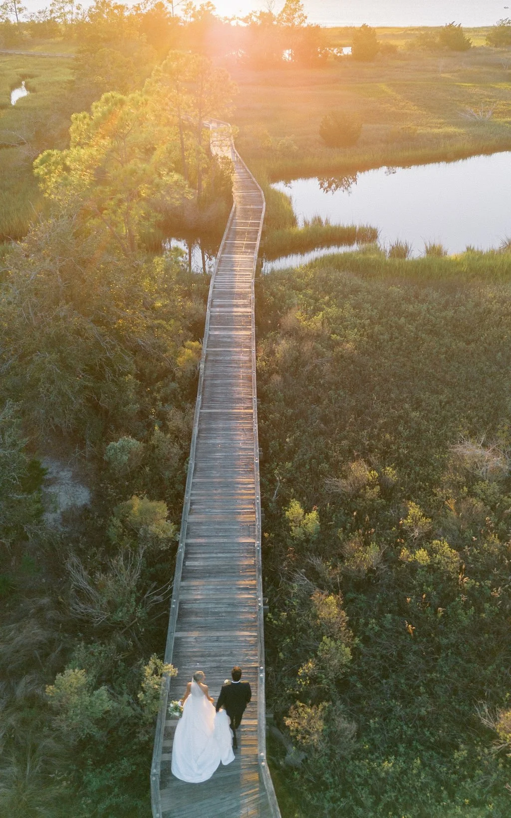 A bride and groom walking hand in hand on a wooden bridge over lush greenery with a pond in the background during sunset.