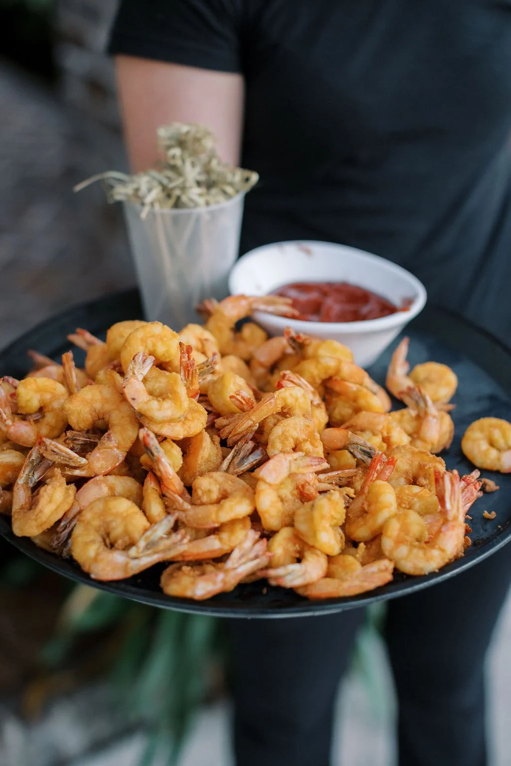 A person holding a black tray of cooked shrimp served with dipping sauce and a side of dried herbs or spices in a plastic cup.