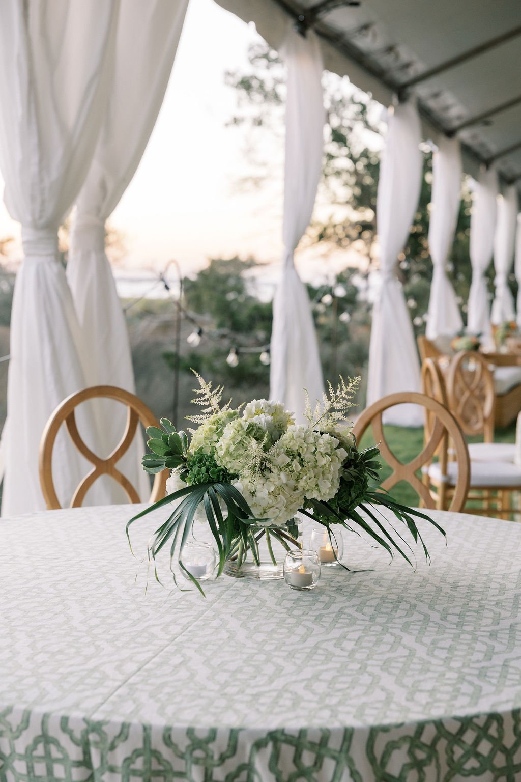 Wedding reception setup with a floral centerpiece on a white tablecloth, surrounded by wooden chairs, and a backdrop of white curtains and trees at sunset.