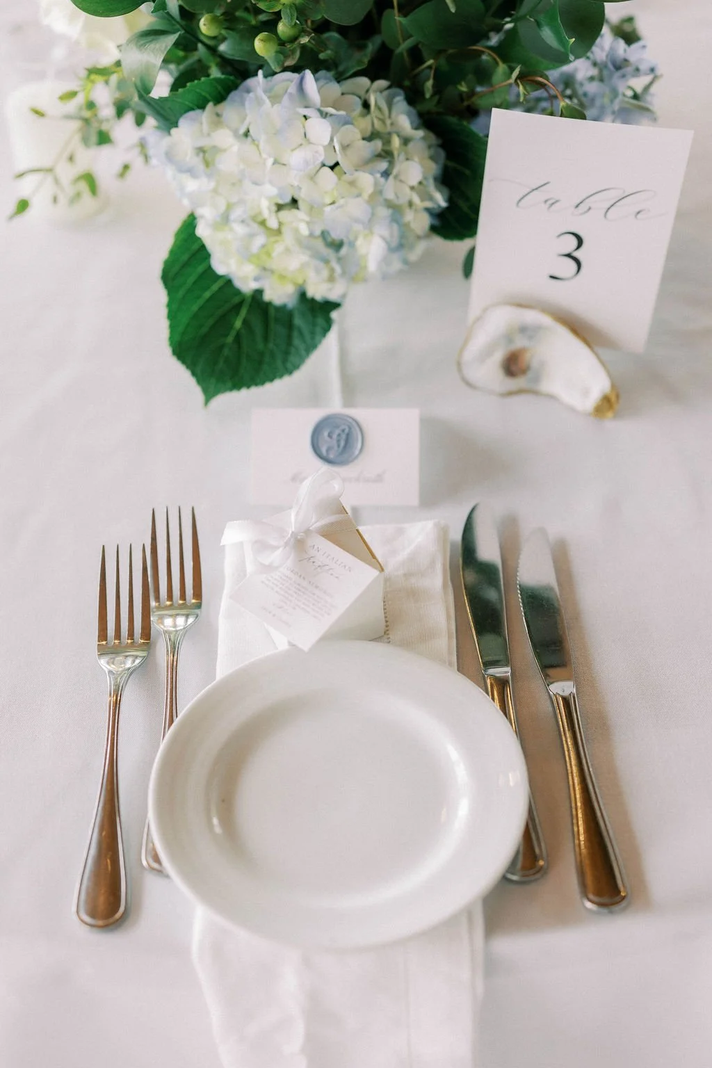 Elegant table setting with a white plate, silver cutlery, a white napkin with a ribboned gift box, a place card, a decorative shell, a flower arrangement with white hydrangeas and green leaves, and a table number card indicating number 3.