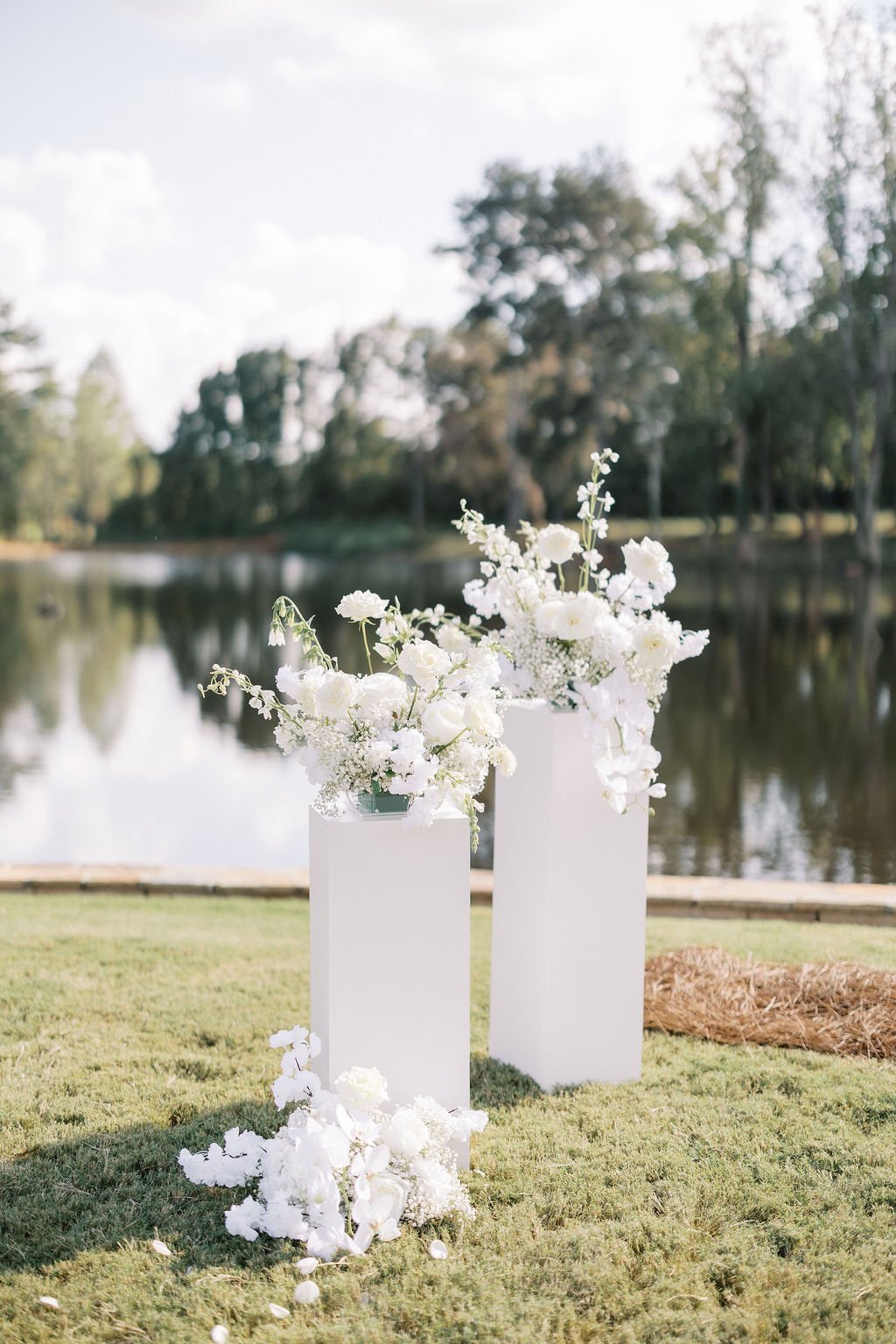 White floral arrangements on two white pedestals by a lake with trees and cloudy sky in the background.
