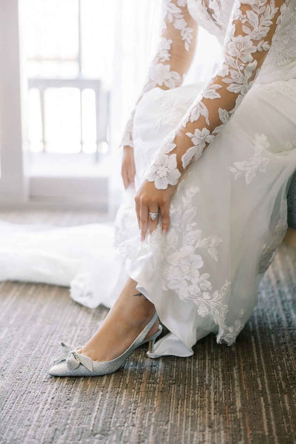 A woman in a white lace wedding dress is sitting on a chair, putting on silver glittery high-heeled shoes with a bow on the toe.