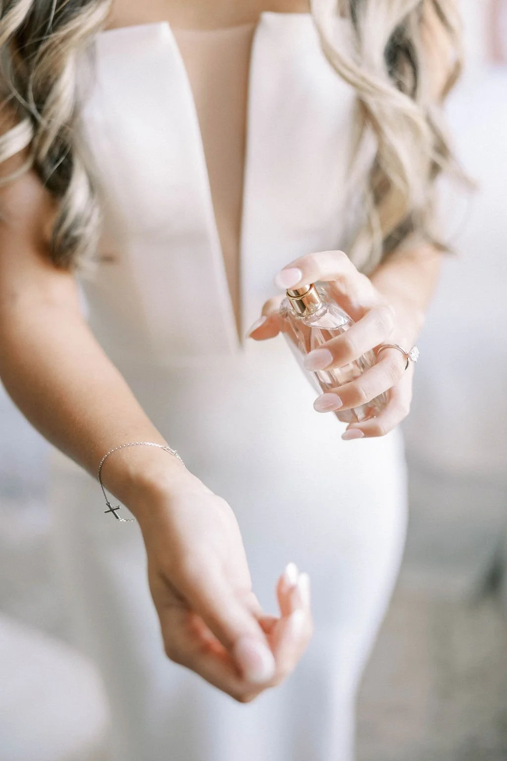A woman in a white dress sprays perfume on her wrist in a softly lit room.
