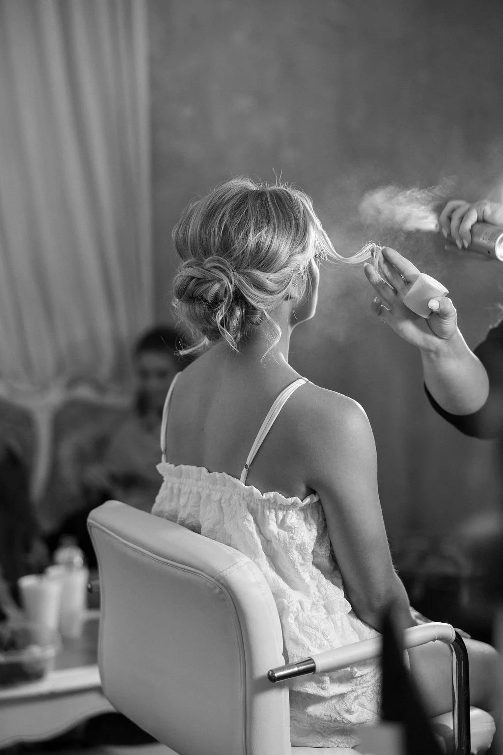A woman getting her hair styled with hair spray at a salon, with a person looking on in the background.