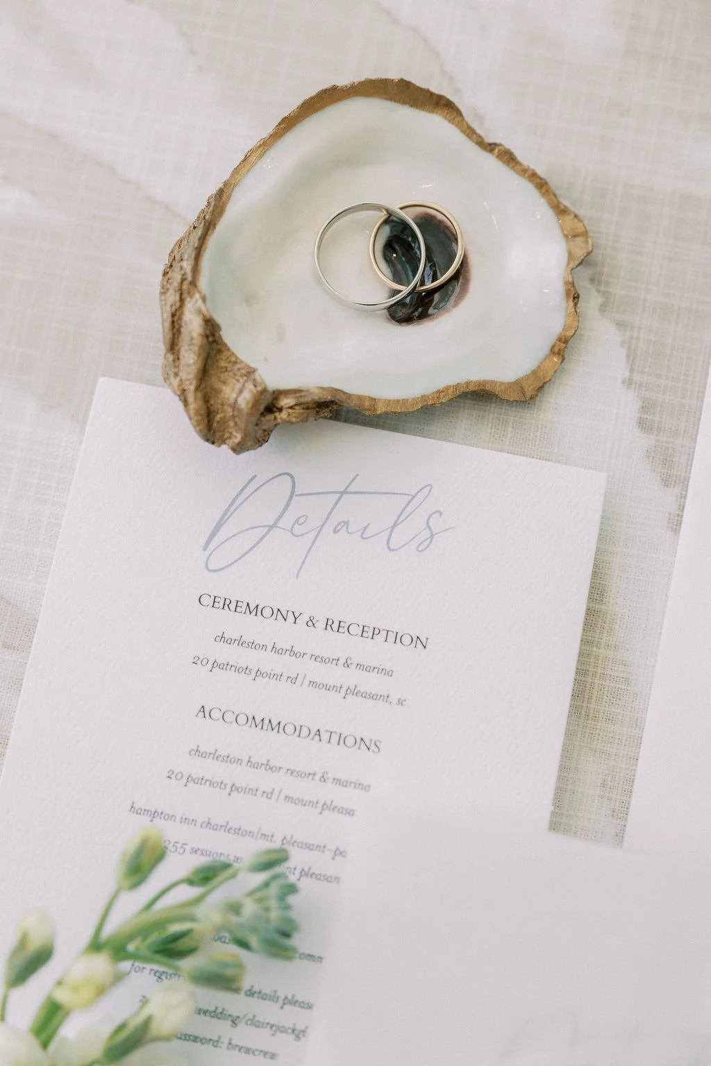 A wedding wedding invitation on white paper with text mentioning details for ceremony and reception at Charleston Harbor Resort & Marina. A large oyster shell holds two rings atop the invitation, and a small white flower arrangement is partially visi