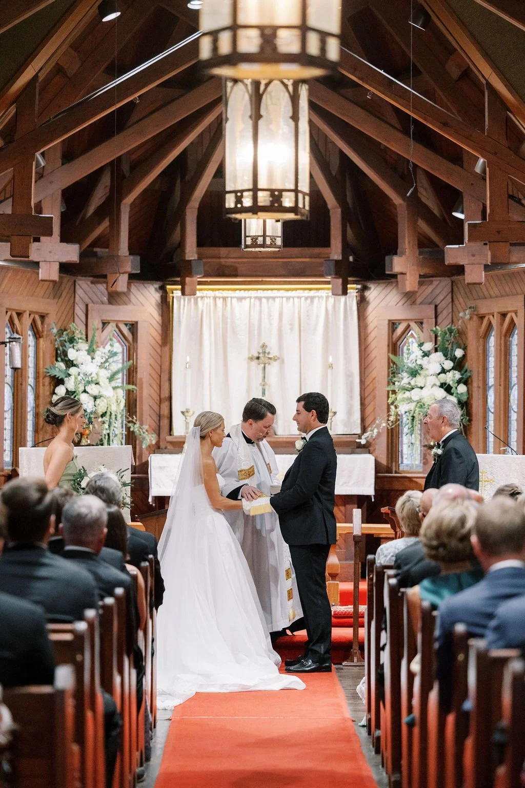 A wedding ceremony inside a wooden church with a bride and groom exchanging vows, facing each other with an officiant, while guests seated in rows watch.