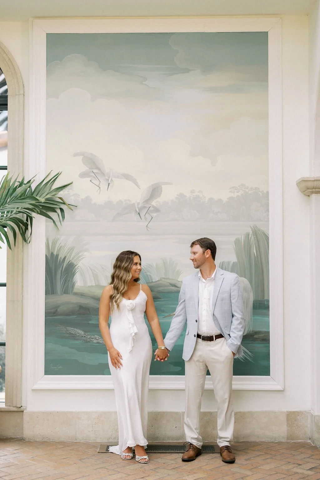 A couple standing together holding hands in front of a painted mural of a river scene with flying herons and trees, indoors with tropical plants nearby.