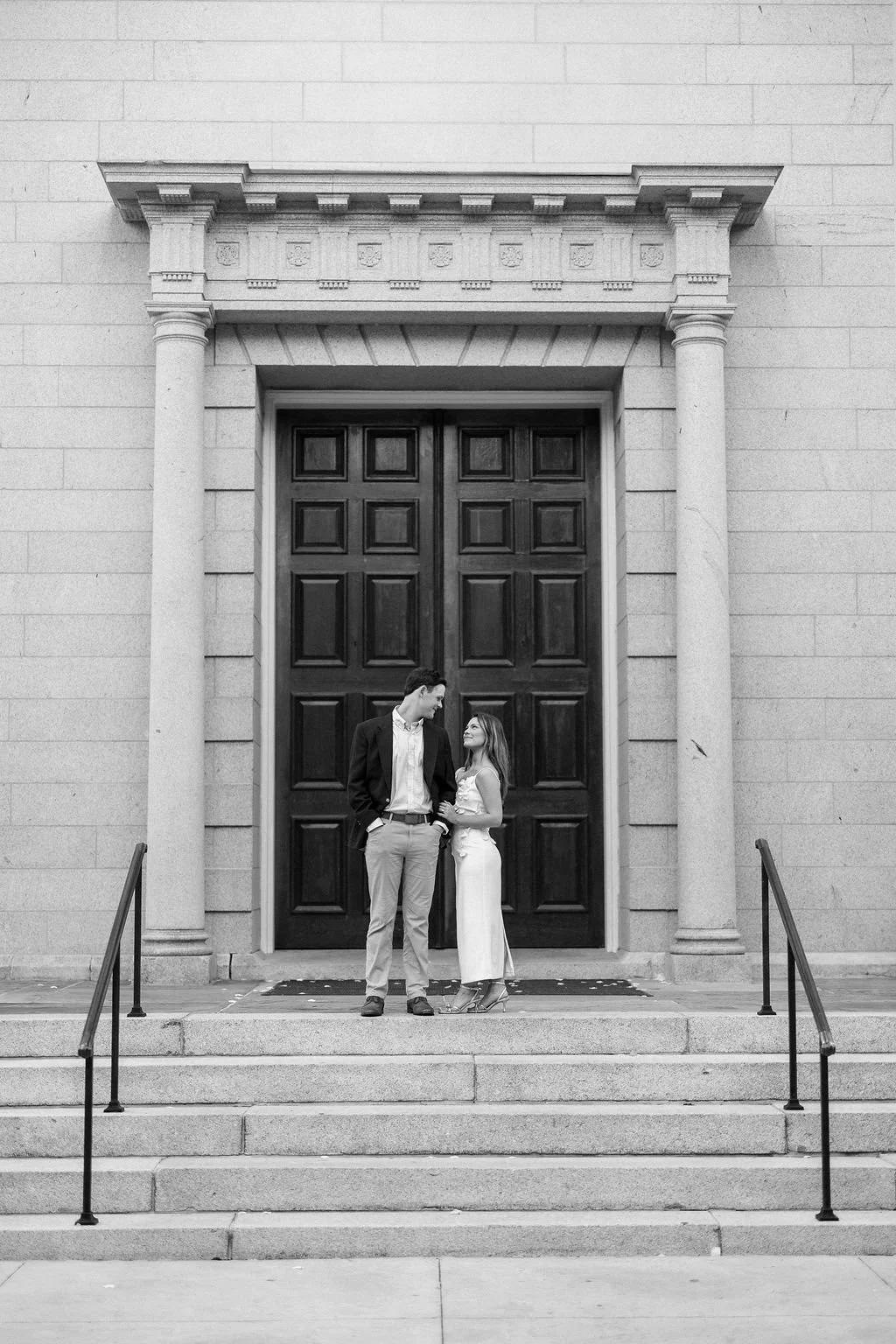 A black and white photo of a couple standing on the steps in front of large wooden doors, smiling and looking at each other. The building has stone columns and decorative architectural details.