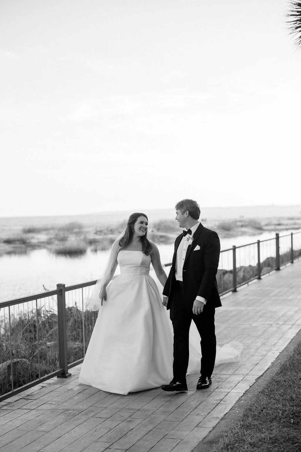 A bride and groom walking hand in hand on a wooden pathway near water, smiling and looking at each other, outdoors during daytime in black and white.