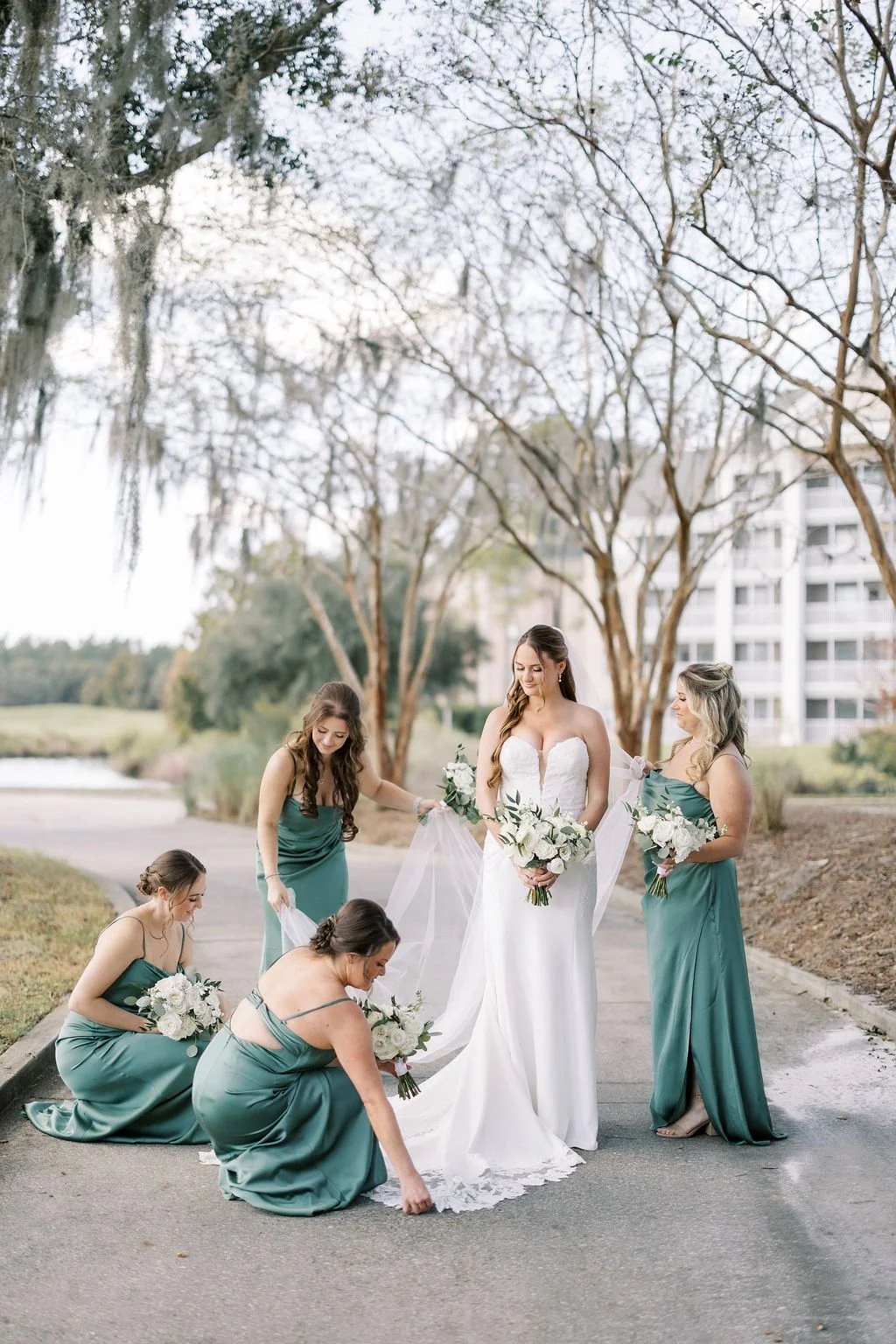 Bride and four bridesmaids in teal dresses posing outdoors, holding bouquets, with trees and a building in the background.