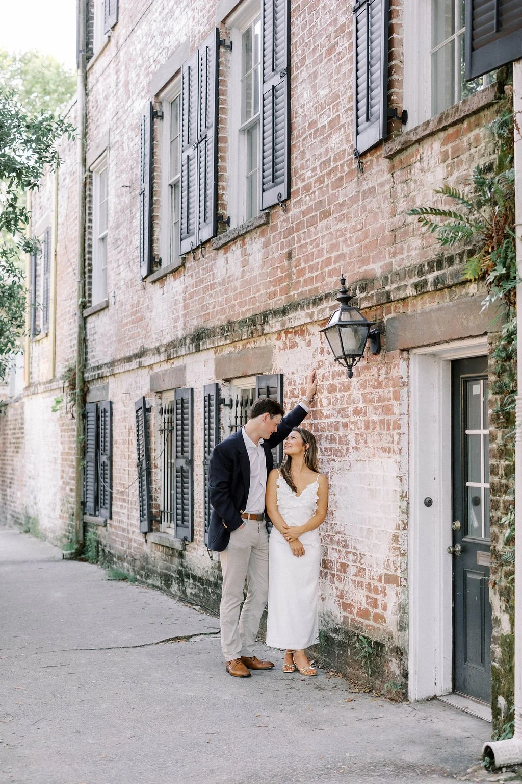 A woman in a white dress and a man in a dark blazer and light pants standing on a city sidewalk, leaning against a brick wall with black shutters, near a black outdoor light fixture, engaging in a romantic moment.