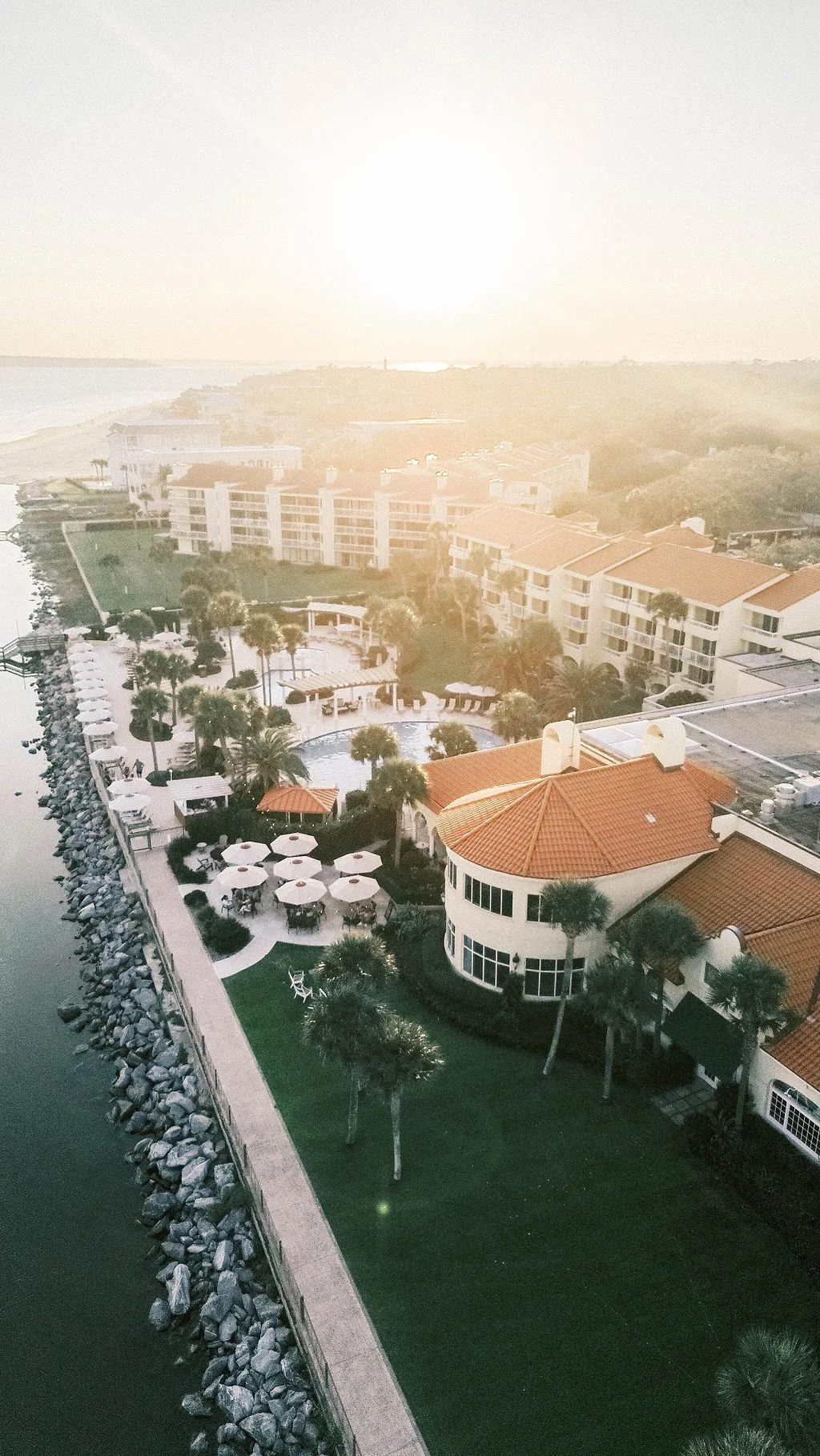 Aerial view of a waterfront resort with a pool, umbrellas, palm trees, and a large building with a red-tiled roof at sunset.