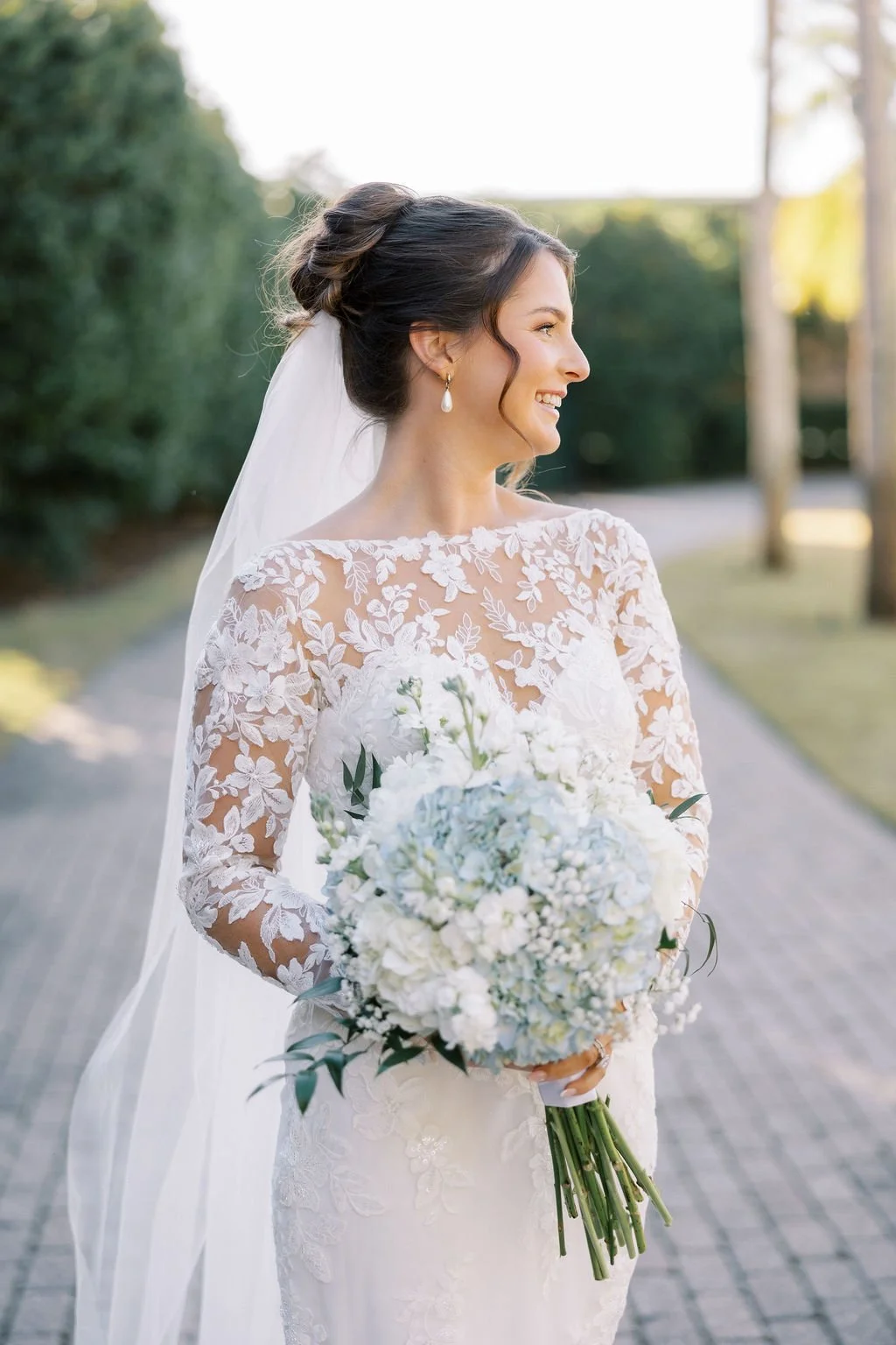 A bride in a white lace wedding dress holding a bouquet of white flowers, standing outdoors on a paved path with greenery and trees in the background.