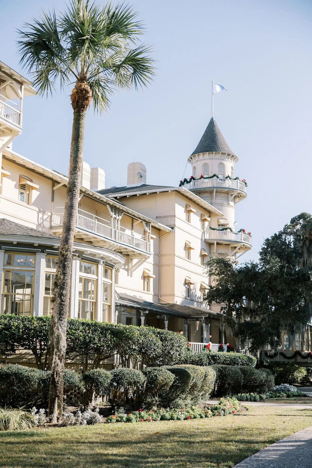 A large beige Victorian-style house with a prominent tower, decorated with holiday garlands, next to a tall palm tree, with a well-manicured lawn and bushes in the foreground.