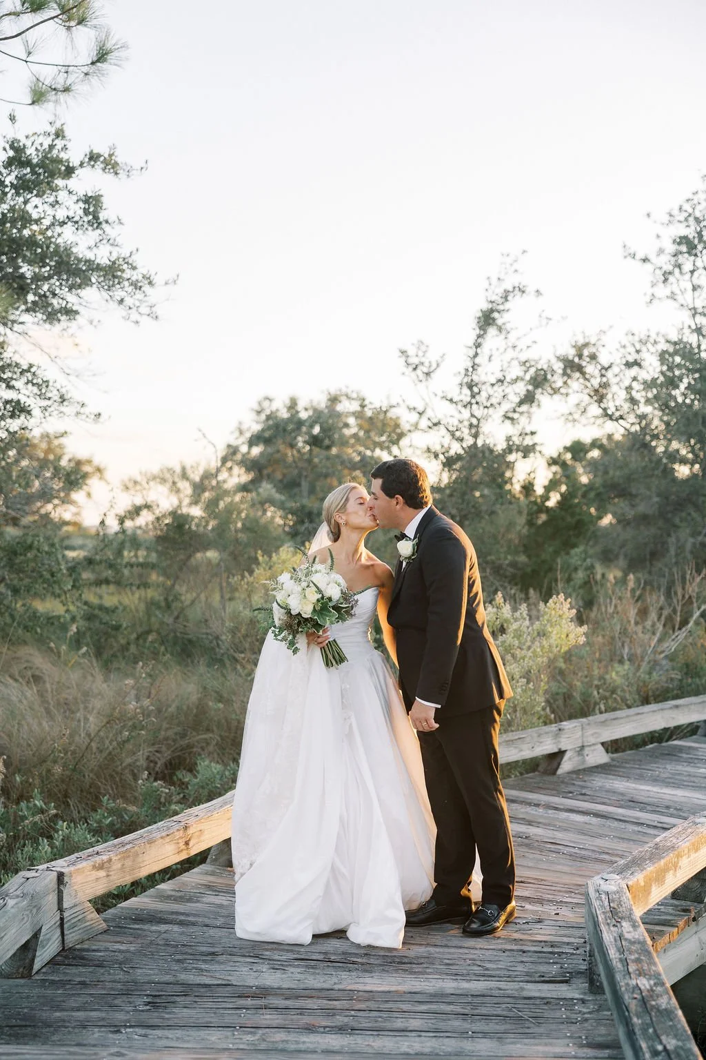A bride and groom sharing a kiss on a wooden bridge outdoors during sunset, with trees and bushes in the background.