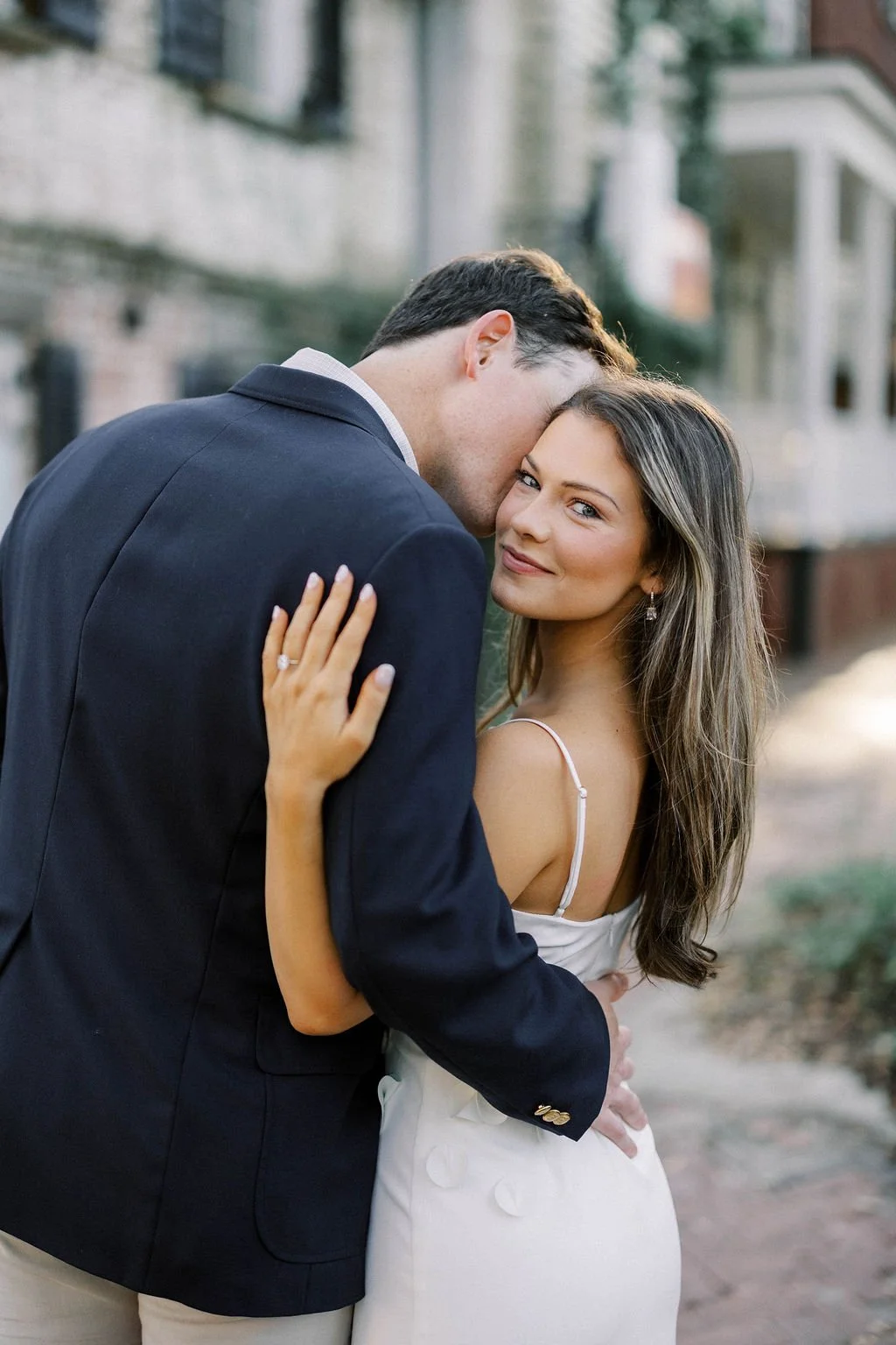 A man and a woman embracing outdoors, with the man resting his forehead on the woman's head, the woman smiling at the camera, wearing a white dress, and the man in a dark blazer.