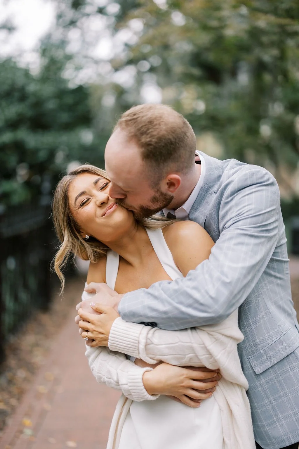 A couple embracing outdoors; the man is kissing the woman on the cheek while she smiles with eyes closed, standing on a path with trees in the background.