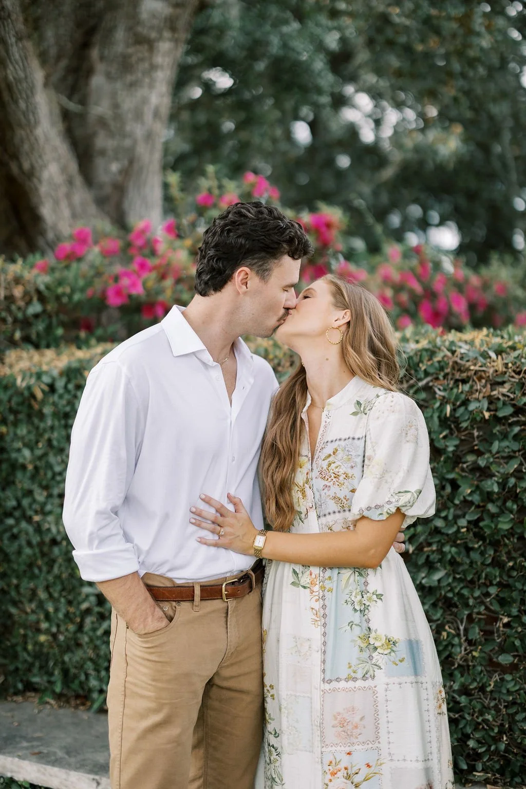 A couple kissing outdoors in front of bushes with pink flowers, a large tree, and greenery.