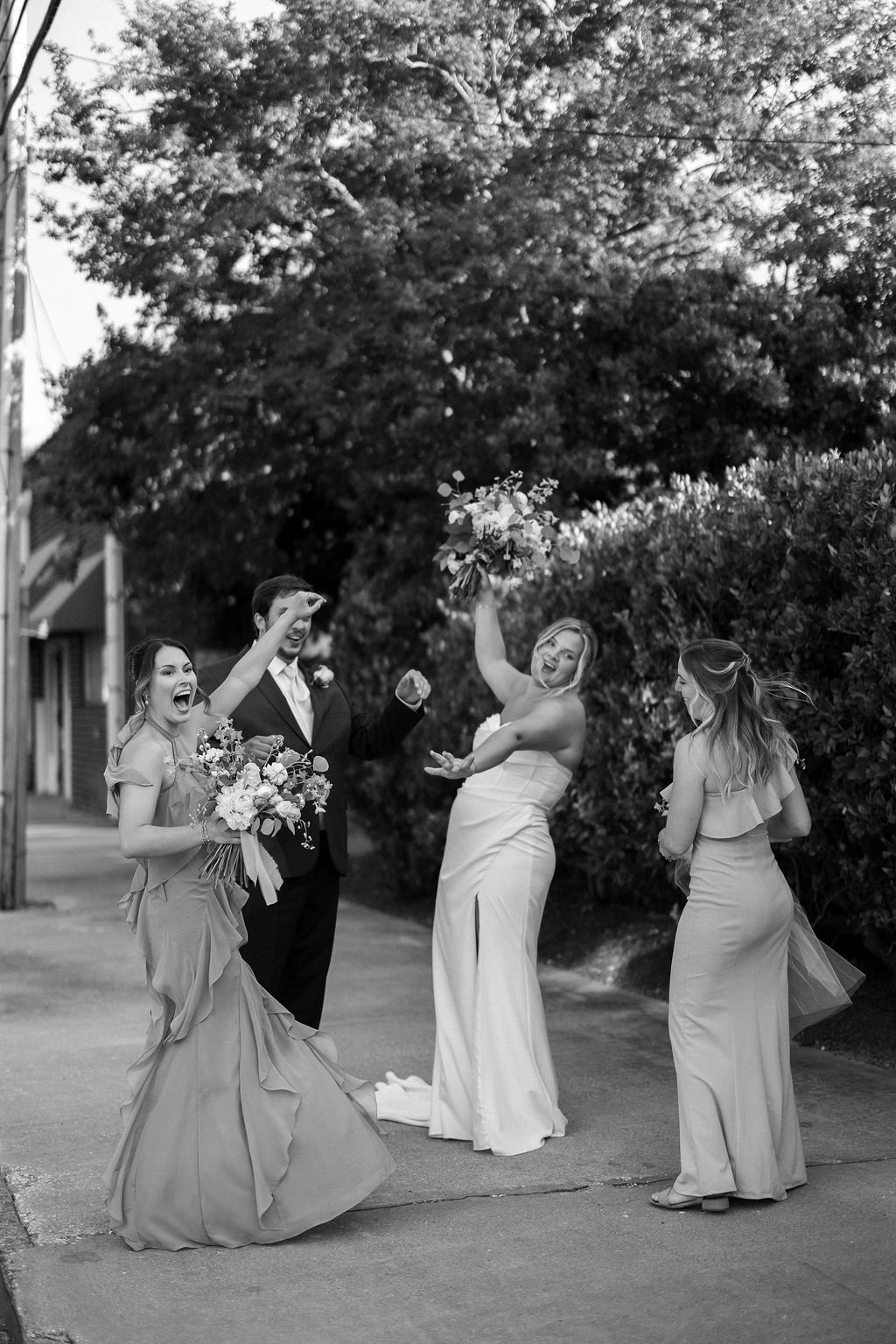 A joyful wedding scene in black and white with four people, including a bride in a wedding dress with a bouquet, a groom in a suit, and two bridesmaids. The bride is tossing a bouquet while the others cheer and laugh on a street lined with trees.