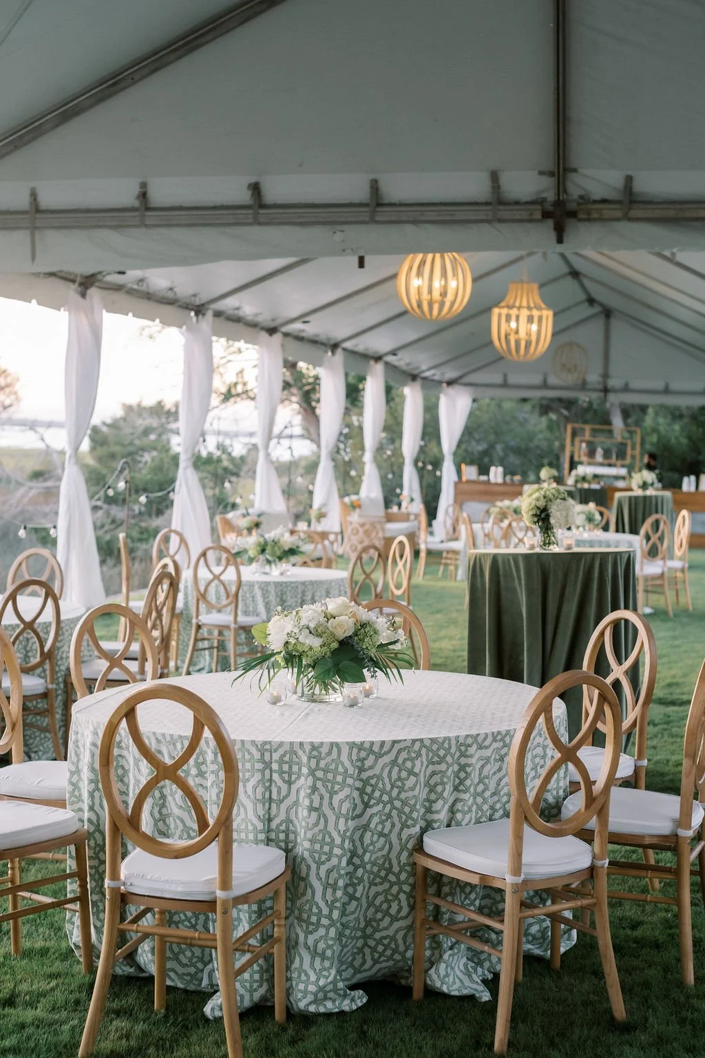 Elegant outdoor event setup with round tables draped in patterned tablecloths, white cushioned chairs, floral centerpieces, and hanging pendant lights under a large tent with white drapes.