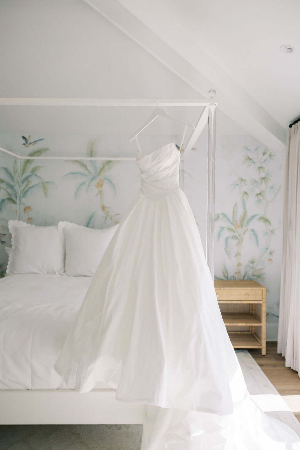 A white wedding dress hanging from a clear canopy in a room with a jungle mural on the wall and a wooden nightstand.