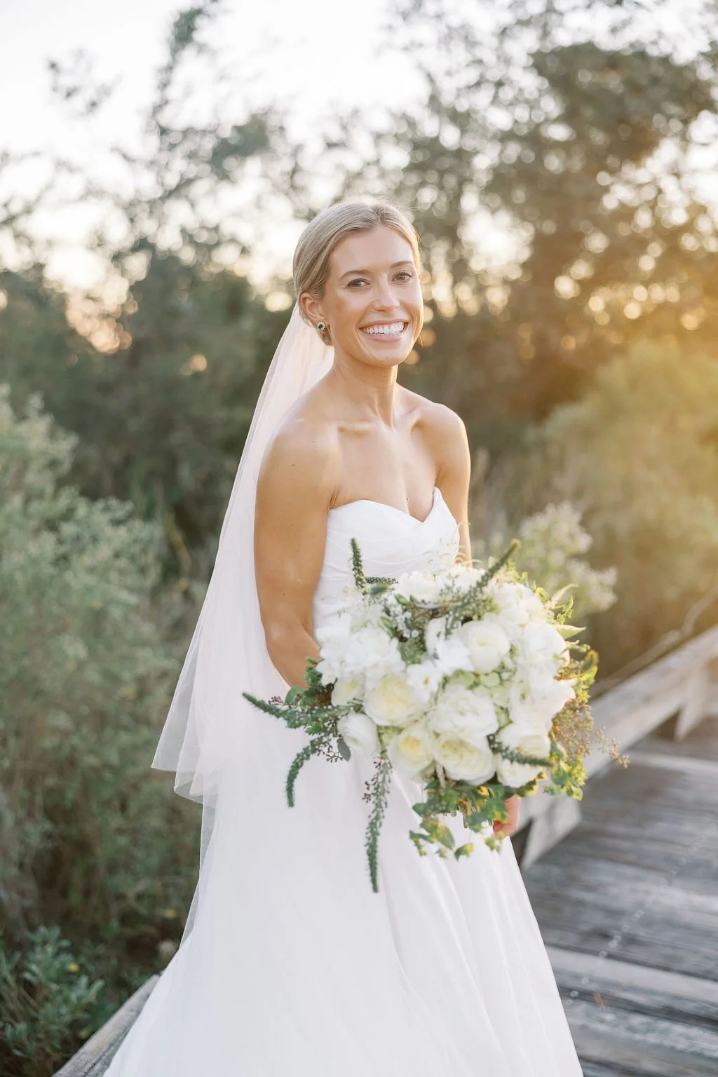 A smiling bride in a strapless white wedding gown holding a large bouquet of white flowers outdoors at sunset.
