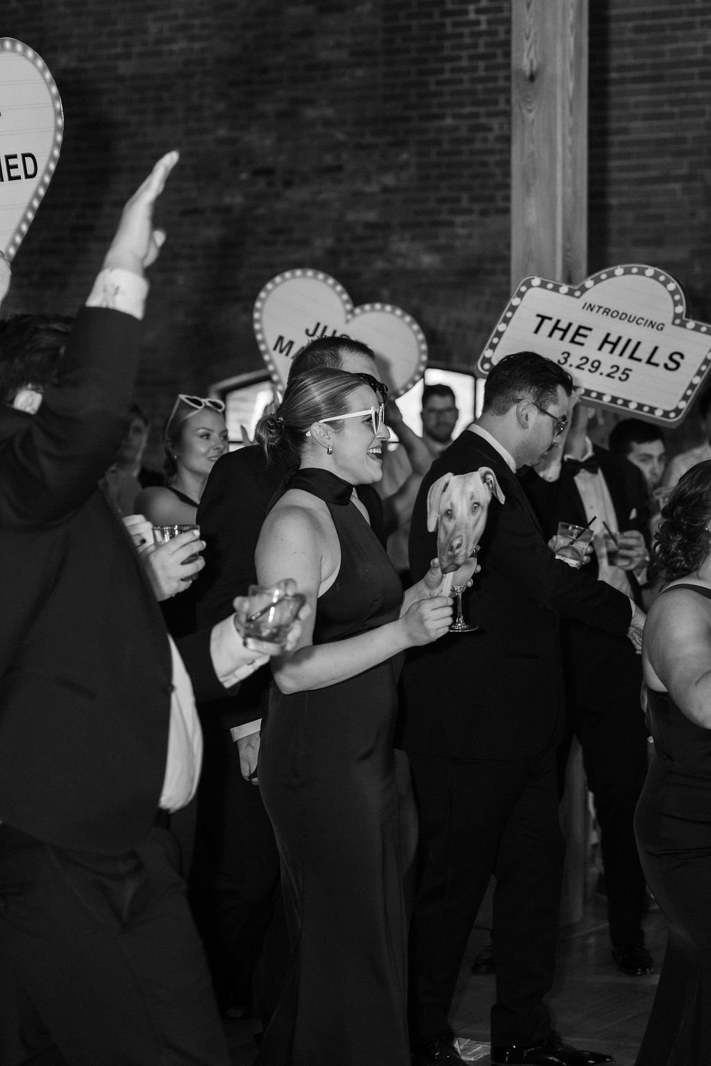 People at a wedding reception, dressed in formal attire, holding drinks, with signs in the background including one that says "Introducing the Hills 3.29.25" and heart-shaped signs with names.