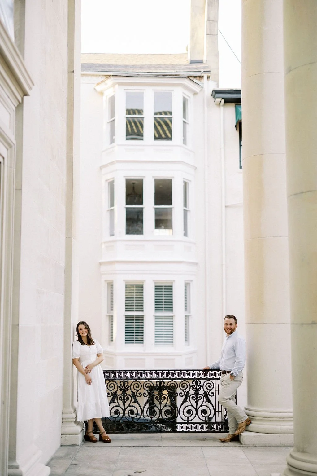 A woman in a white dress and a man in a white shirt and beige pants standing near a decorative black wrought iron railing in a tall, classic building with cream-colored walls and large windows.
