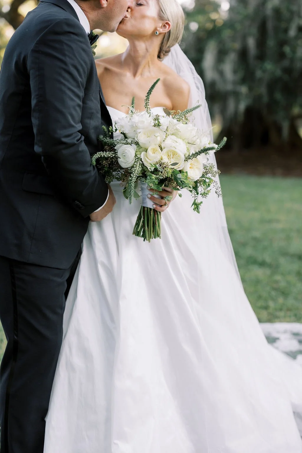 A bride and groom kissing outdoors, with the bride holding a bouquet of white flowers and greenery.